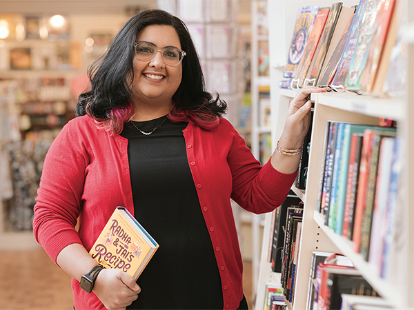 A woman in a black shirt and red cardigan holds a book in one hand as she stands besides a full bookstore shelf.